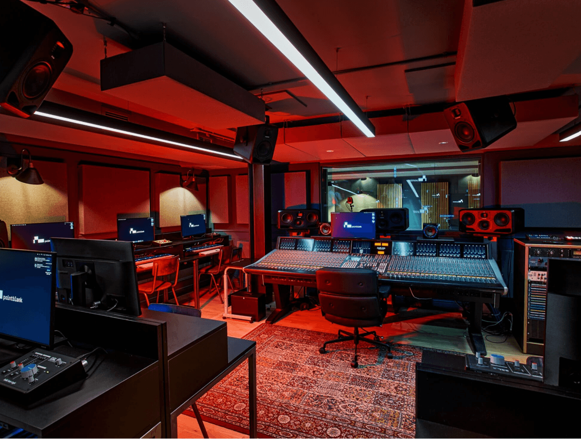 Professional music studio with a large mixing console, red lighting, and multiple computer monitors.
