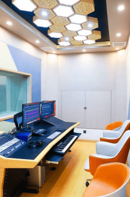 Professional recording studio with dual monitors, keyboard, orange chairs, and hexagonal ceiling light panels.